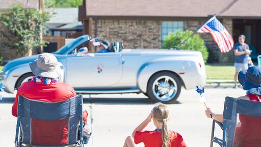 Panorama Caucasian family on camping chairs, sun hats watching car parade on residential street of Coppell, Texas, Independence Day Freedom celebration, utility wagon cart, picnic mat, pinwheels
