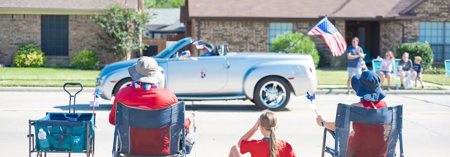 Panorama Caucasian family on camping chairs, sun hats watching car parade on residential street of Coppell, Texas, Independence Day Freedom celebration, utility wagon cart, picnic mat, pinwheels