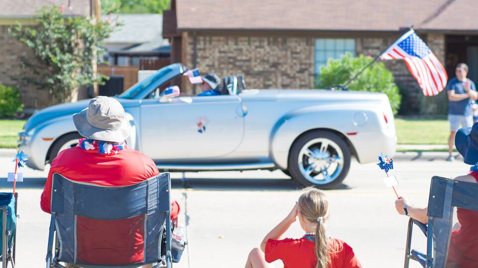 A vintage convertible car, two people in wheelchairs, and a residential street with houses.