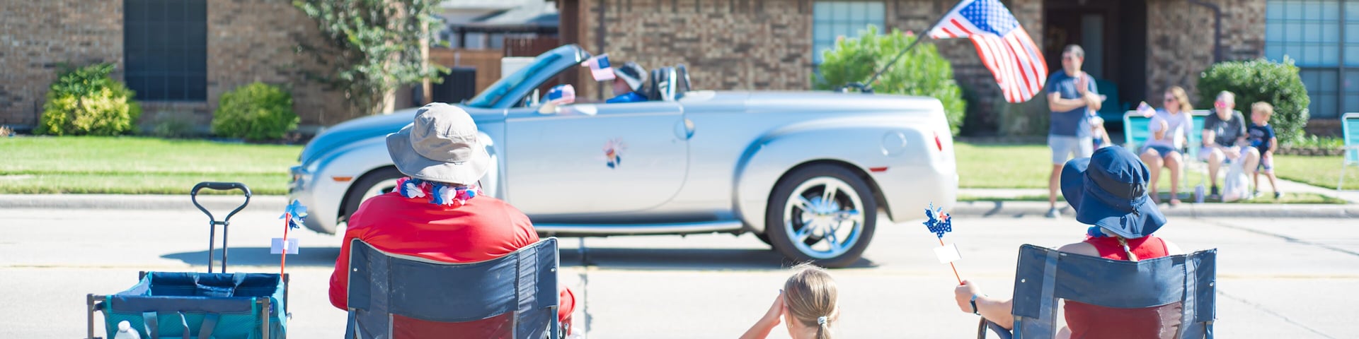 Panorama Caucasian family on camping chairs, sun hats watching car parade on residential street of Coppell, Texas, Independence Day Freedom celebration, utility wagon cart, picnic mat, pinwheels