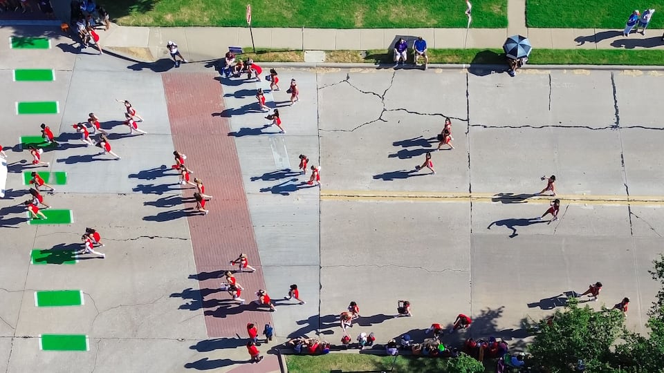 Vista aérea de un desfile con participantes en uniformes rojos, colchonetas verdes y un vehículo azul.