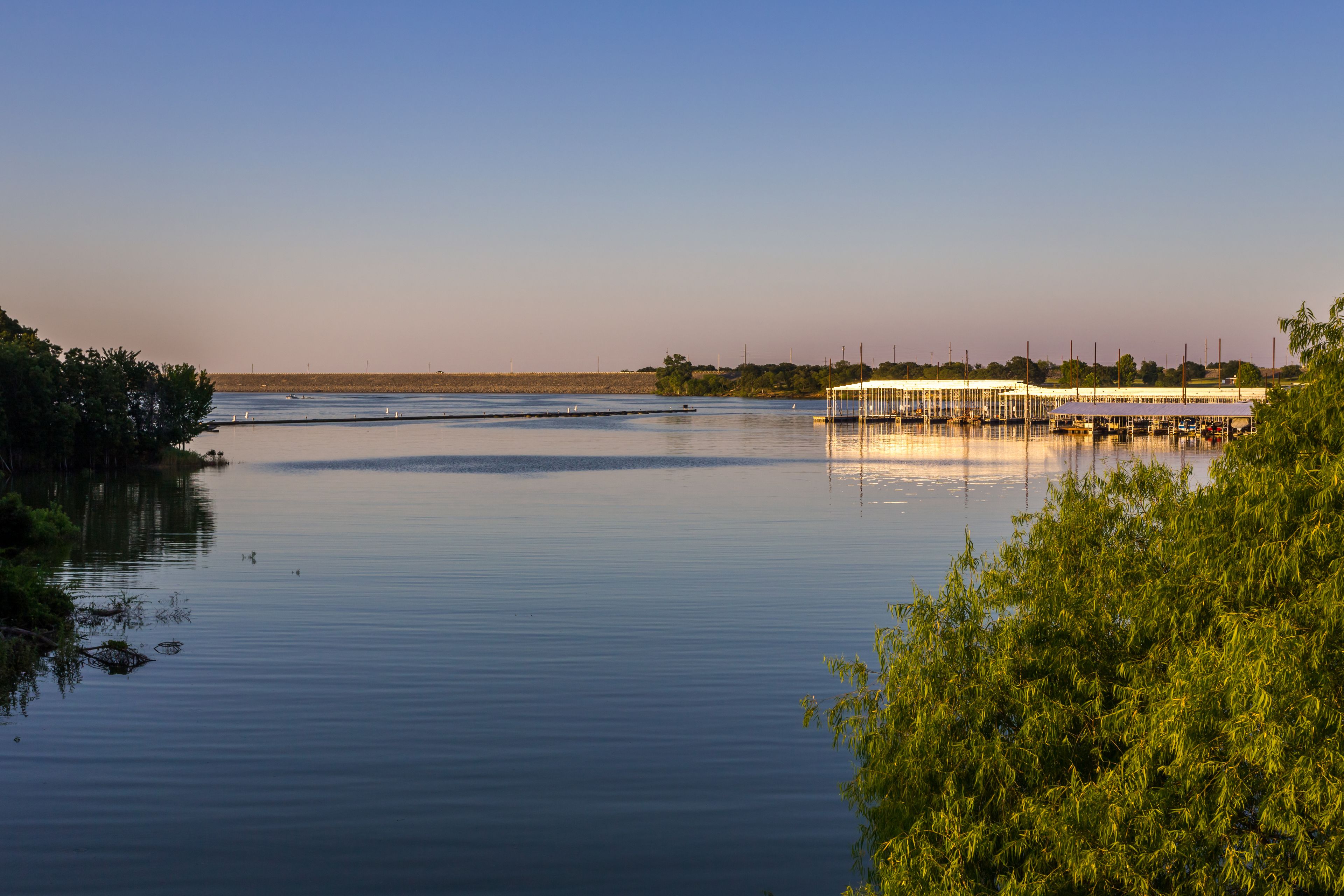 A calm river with a dock and boats, surrounded by greenery and a clear sky.