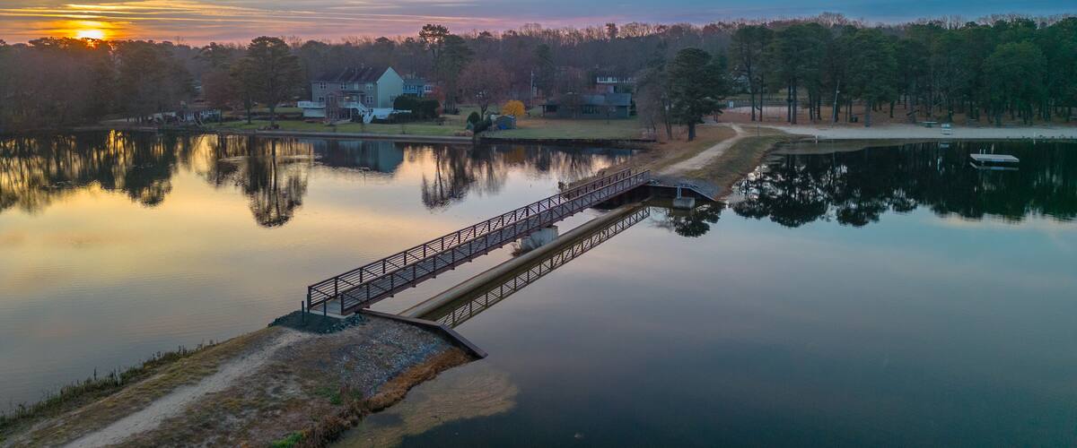 A tranquil lake with a wooden bridge, houses, and trees in the background.
