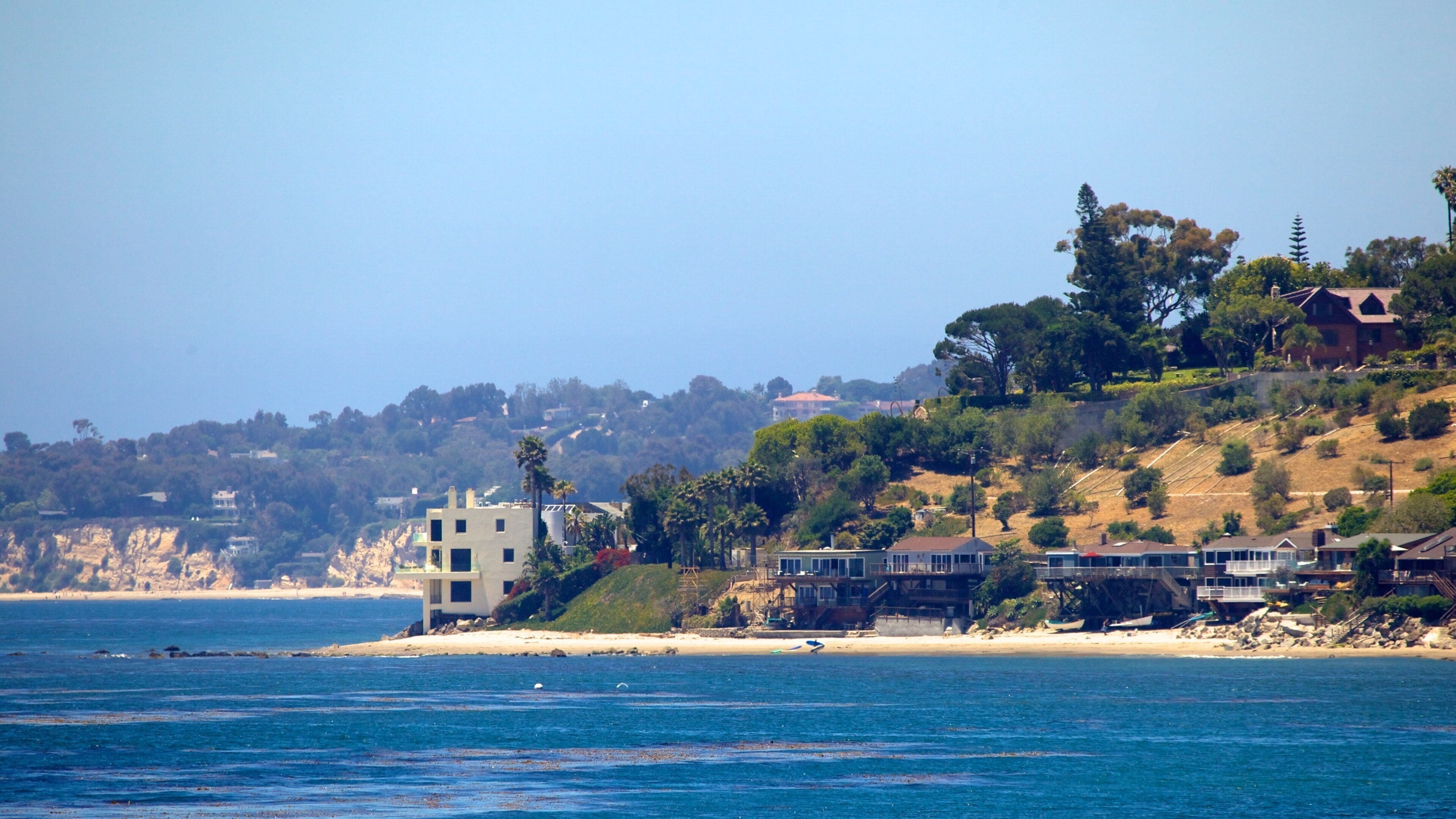 A coastal landscape with a sandy beach, a large body of water, and a hillside with residential buildings and trees.