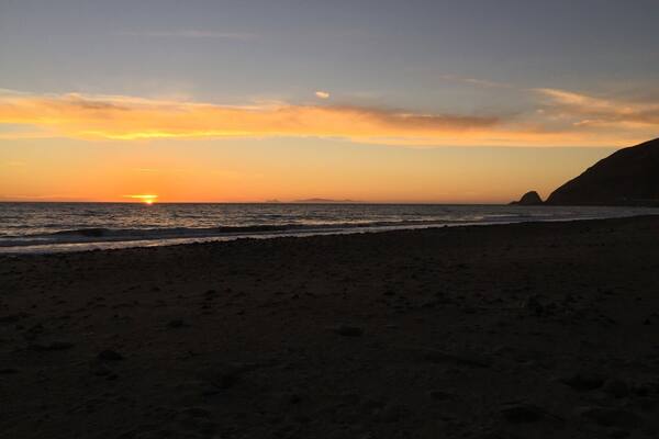 A beach at sunset with the sun near the horizon, waves, and a distant mountain.