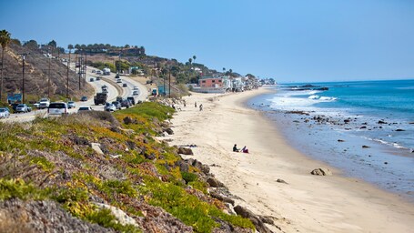 A coastal road with vehicles, a beach, and buildings on a hillside.