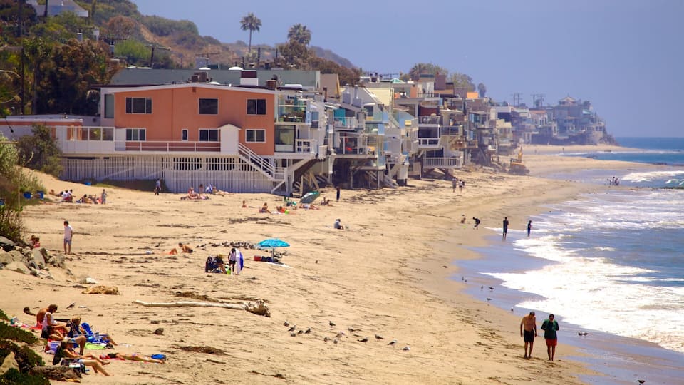 A beach with people sunbathing and walking, a lifeguard tower, and houses built on stilts near the water.