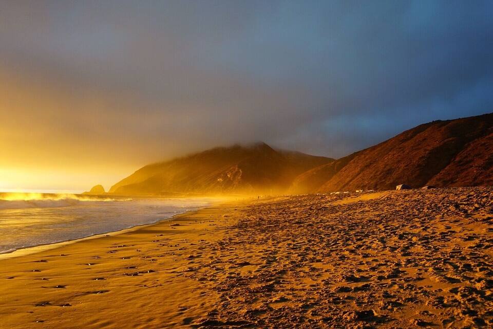 A beach with golden sand and waves crashing onto the shore, with a mountain in the background.