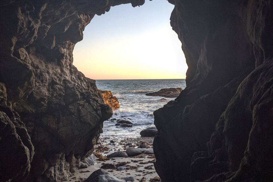 A rocky cave opening to a beach with waves and a sunset.