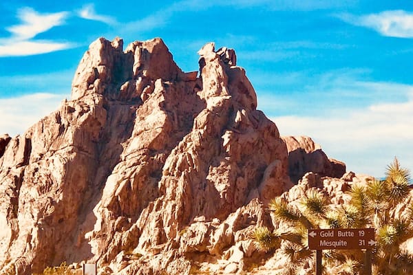 A rocky mountain with a clear blue sky. There is a signpost with directions to 'Gold Butte 19' and 'Mesquite 31'.