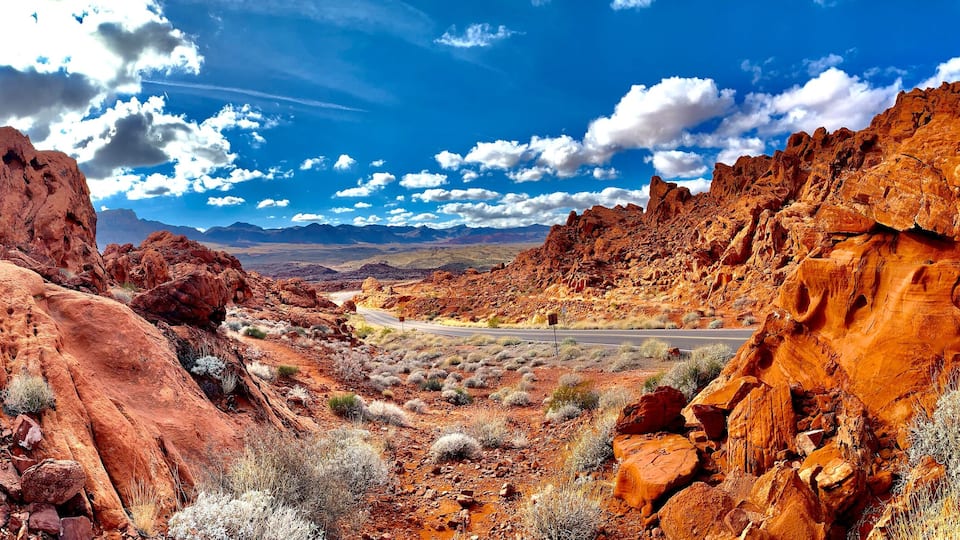 A desert landscape with red rock formations, sparse vegetation, and a clear blue sky with clouds.