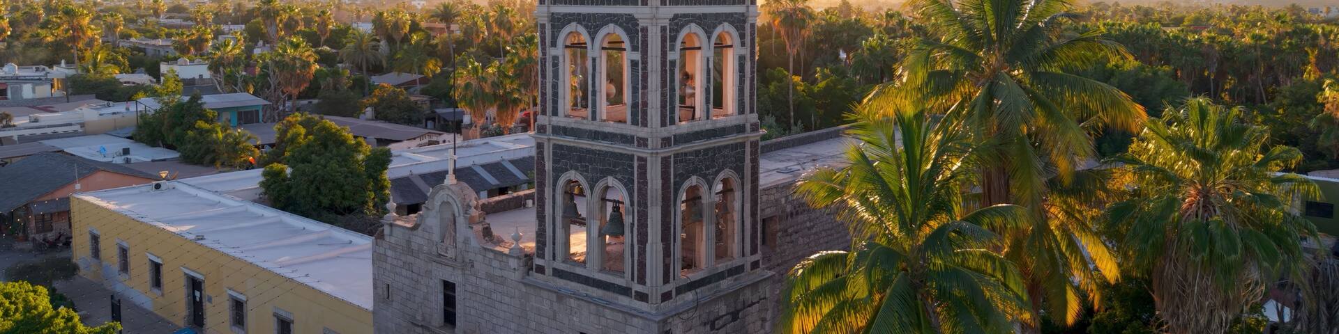 A historic church with a bell tower, palm trees, and mountains in the background.