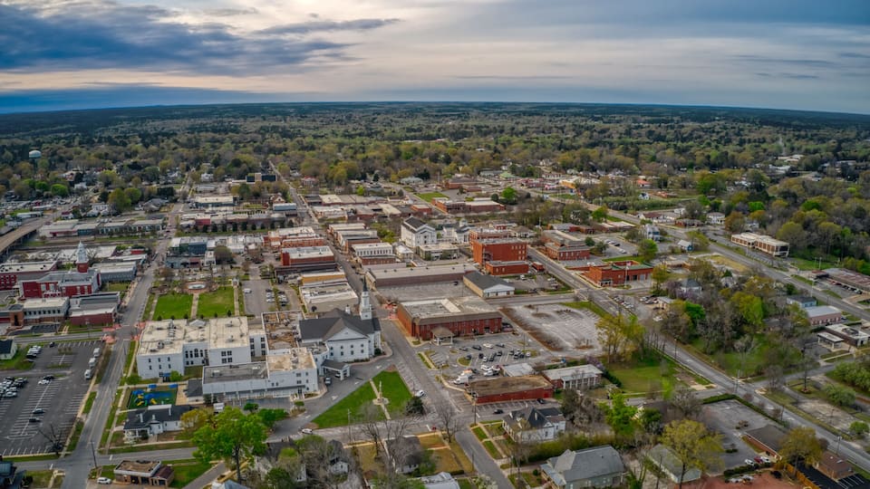 An aerial view of a town with a mix of commercial and residential buildings, a prominent church, and surrounding greenery.