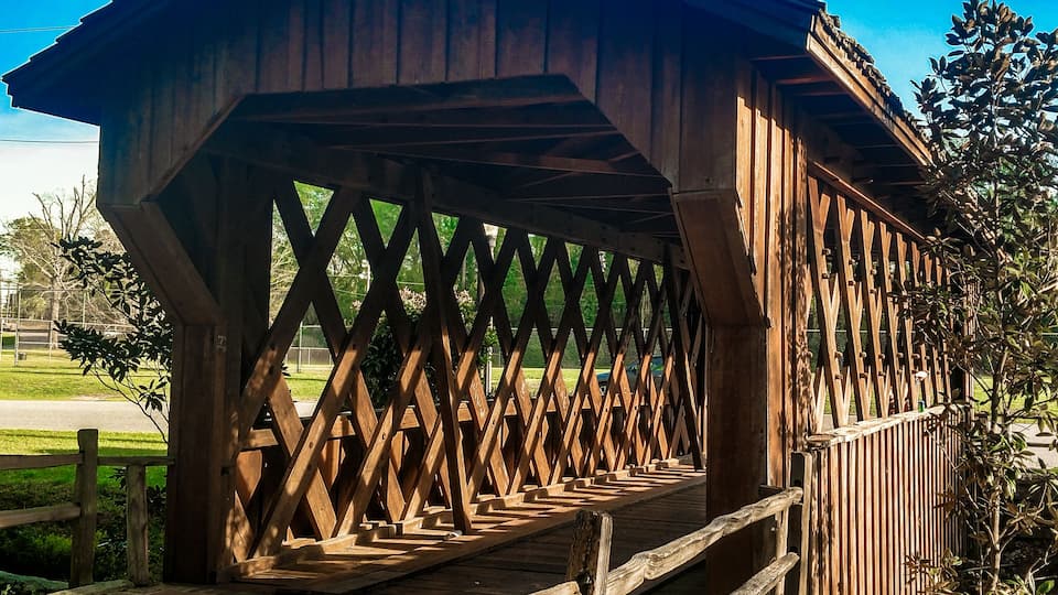 A wooden covered bridge with a lattice design, surrounded by greenery and a clear blue sky.