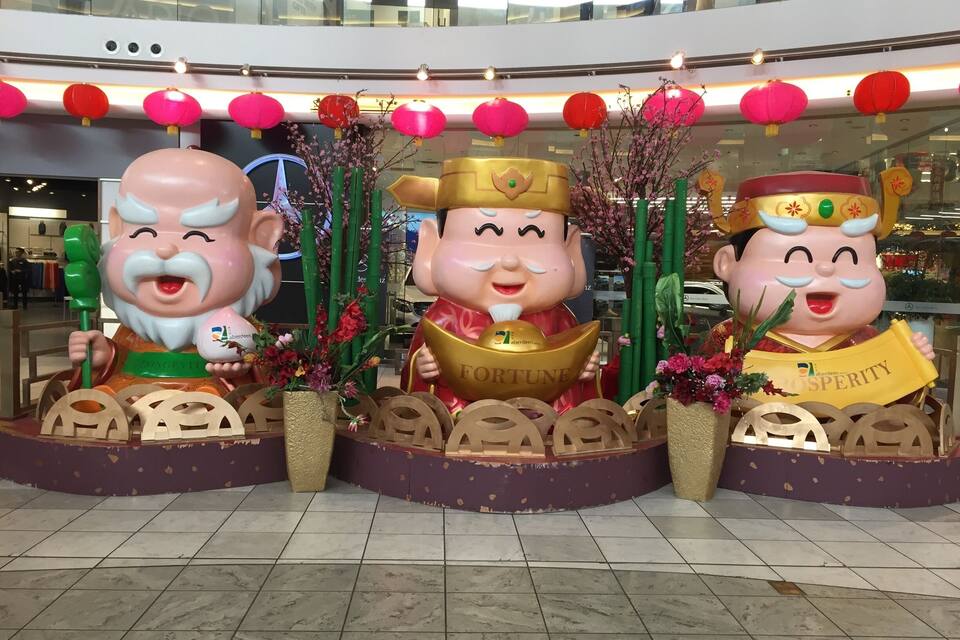 Exposição decorativa com três figuras imponentes segurando objetos, cercada por lanternas vermelhas e arranjos florais.