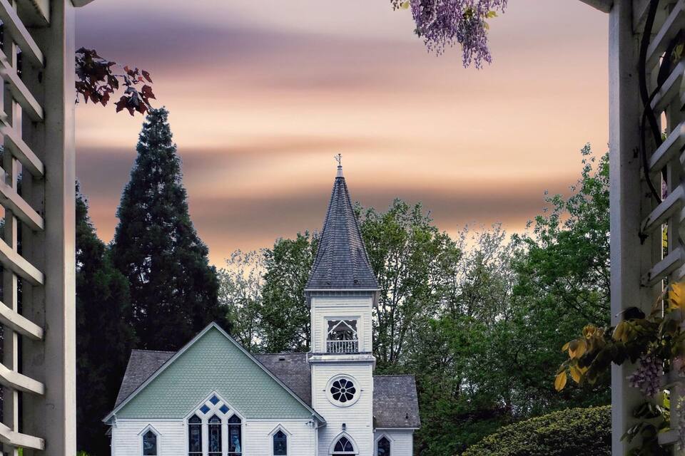 Igreja com um campanário e torre do relógio, rodeada por vegetação e um arco de madeira.