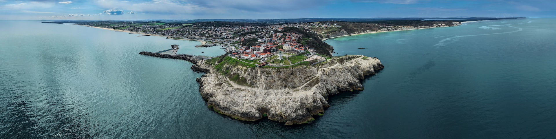 An aerial view of a coastal town with a prominent rocky island, a lighthouse, and a clear view of the surrounding sea and distant shoreline.
