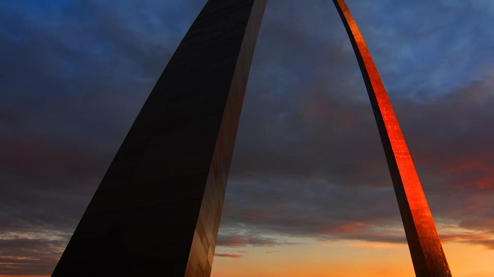 A large arch structure with a red illuminated section against a twilight sky.