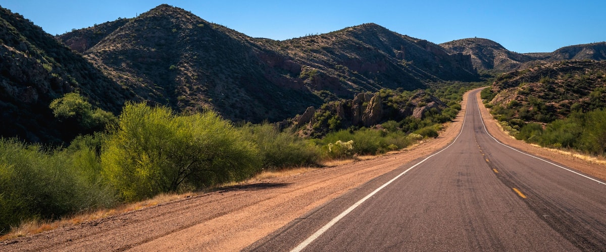 A straight road flanked by green vegetation and rocky mountains under a clear blue sky.