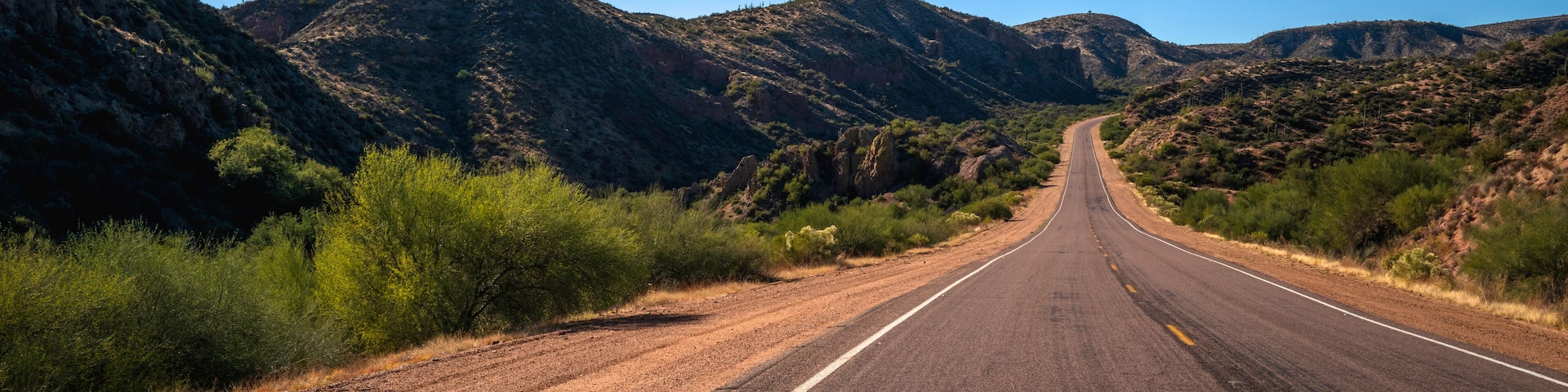 A straight road flanked by green vegetation and rocky mountains under a clear blue sky.