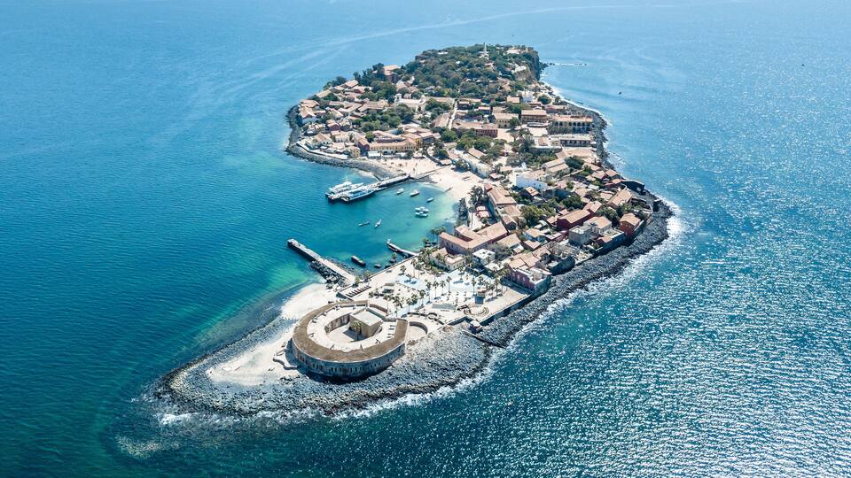 An aerial view of a coastal town with a fortified structure, a harbor with boats, and a clear blue sea.