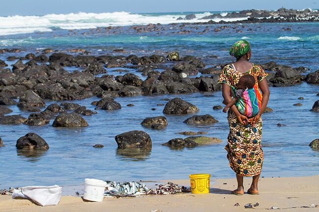 Uma mulher está em pé numa praia rochosa com uma criança, segurando um tecido colorido. Há dois baldes e uma bolsa branca na areia.