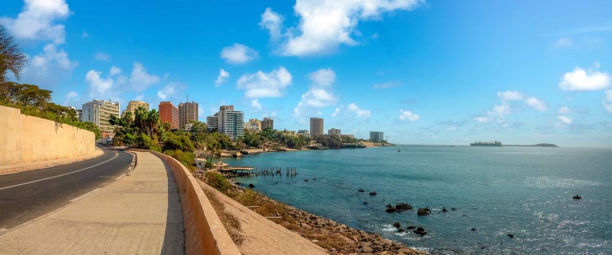 A coastal road with a city skyline in the background, a clear blue sky, and calm sea.