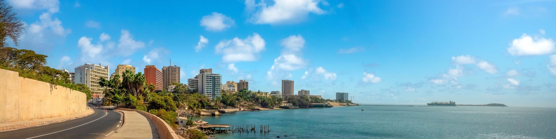 A coastal road with a city skyline in the background, a clear blue sky, and calm sea.