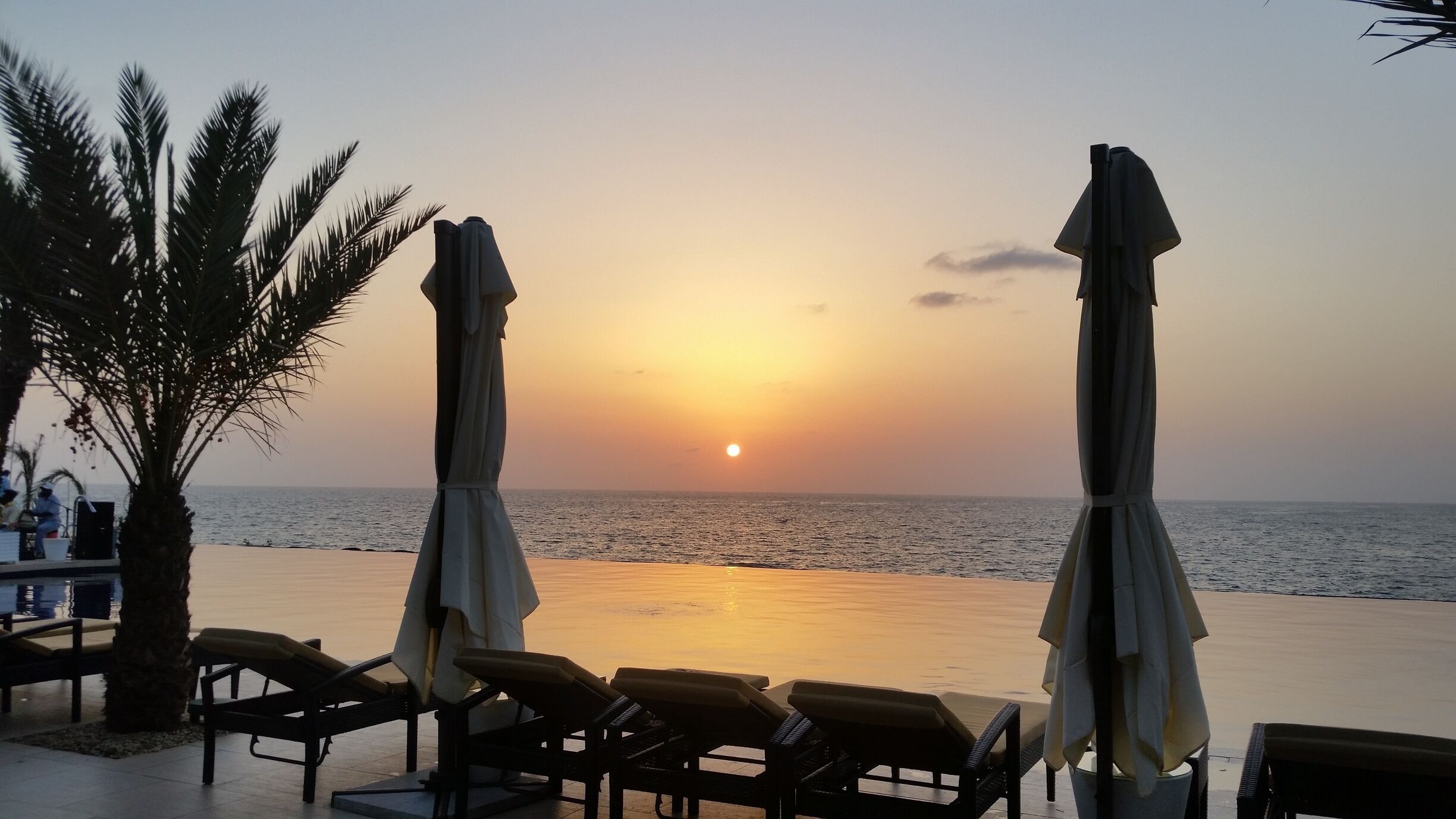 A beach with lounge chairs and umbrellas, palm trees, and a sunset over the ocean.