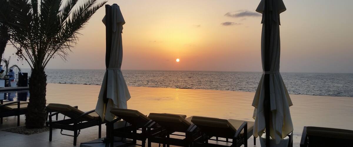 A beach with lounge chairs and umbrellas, palm trees, and a sunset over the ocean.