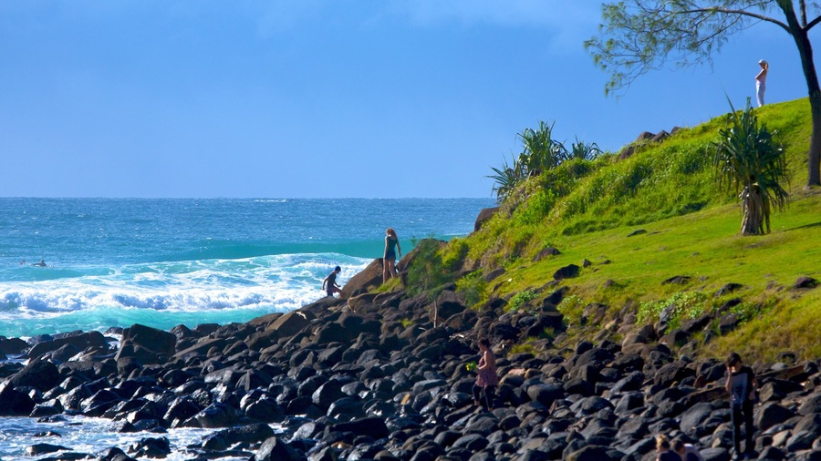 A rocky shoreline with waves crashing onto the rocks, a grassy hill with people standing on it, and a clear blue sky.