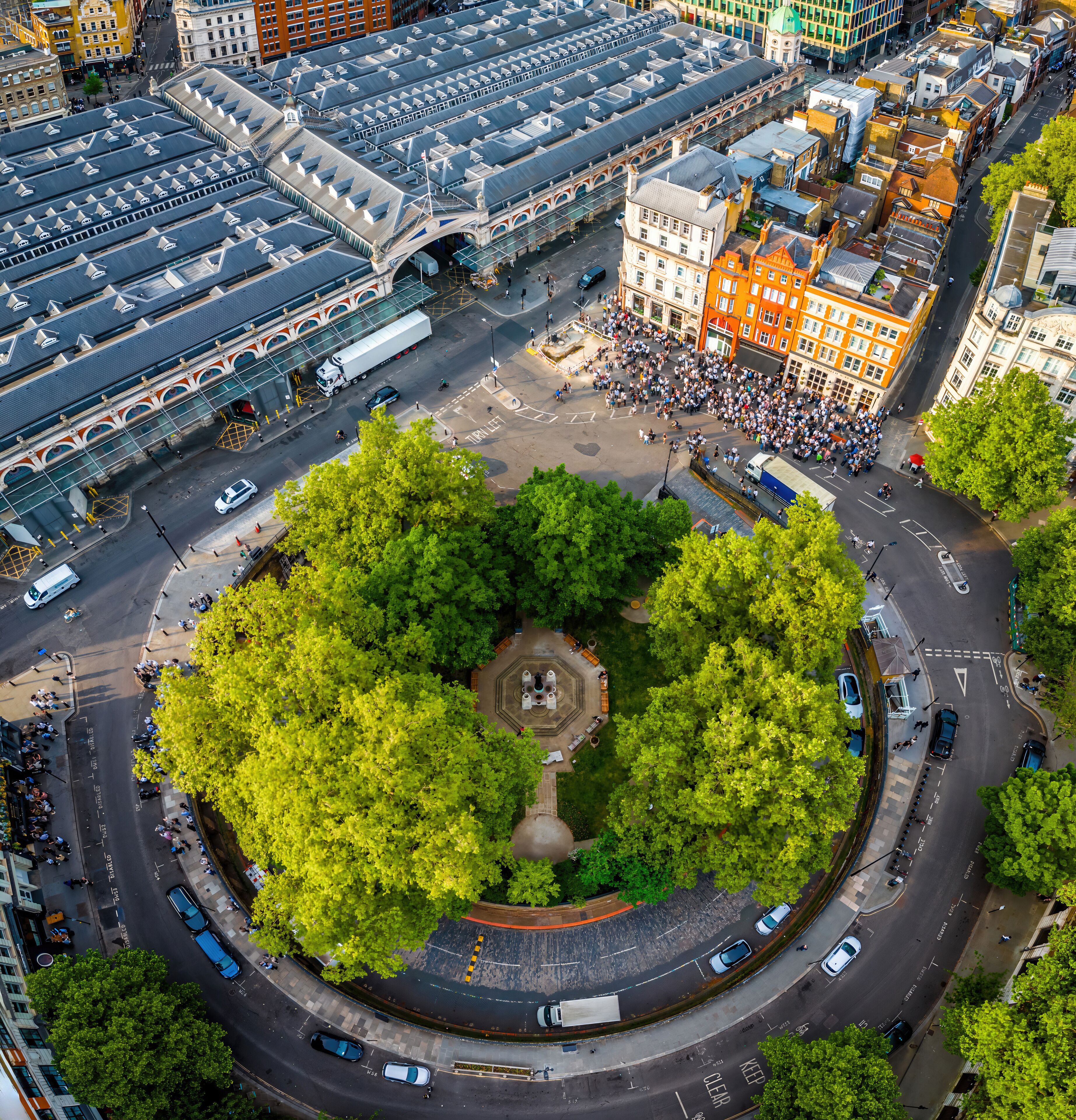 A roundabout with a central green space surrounded by trees, a historic building with a clock tower, and a tram station with a roof.