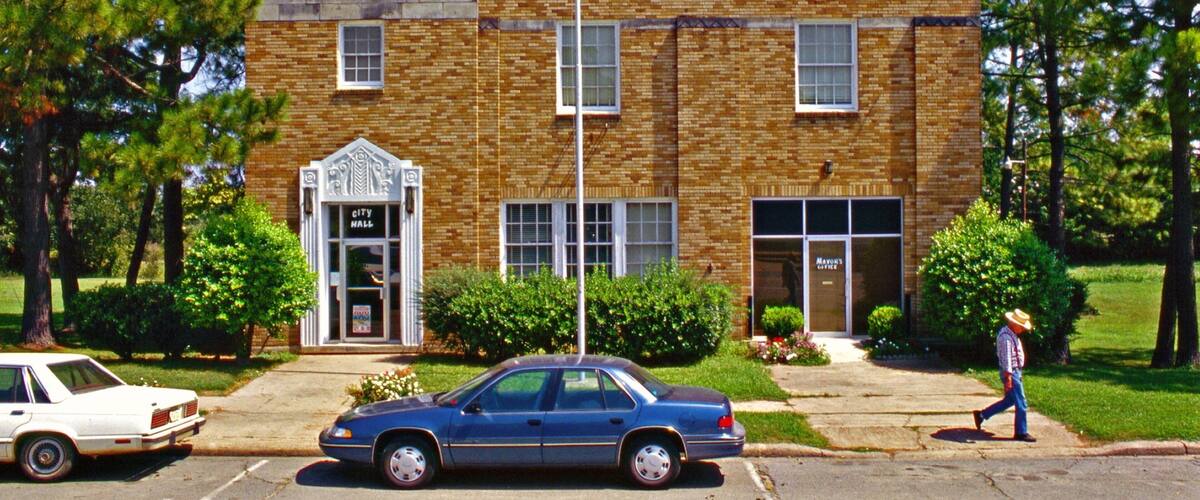 A city hall building with a brick facade, a blue car parked in front, and a person walking on the sidewalk.