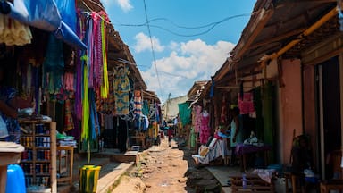 A narrow street with colorful textiles hanging from stalls, wooden buildings, and a clear blue sky.