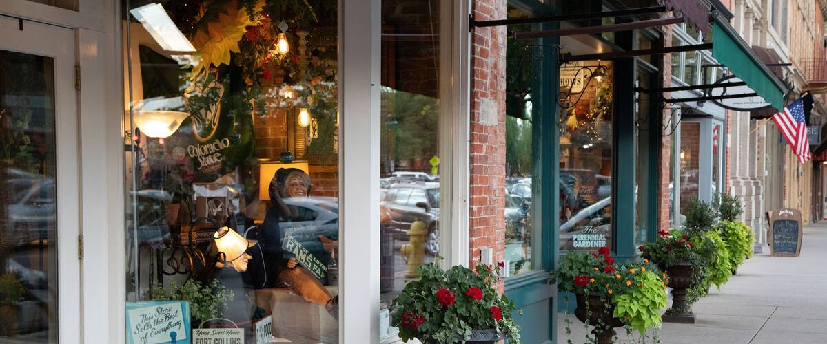A storefront with a large window display, a brick building, and a row of potted plants on the sidewalk.