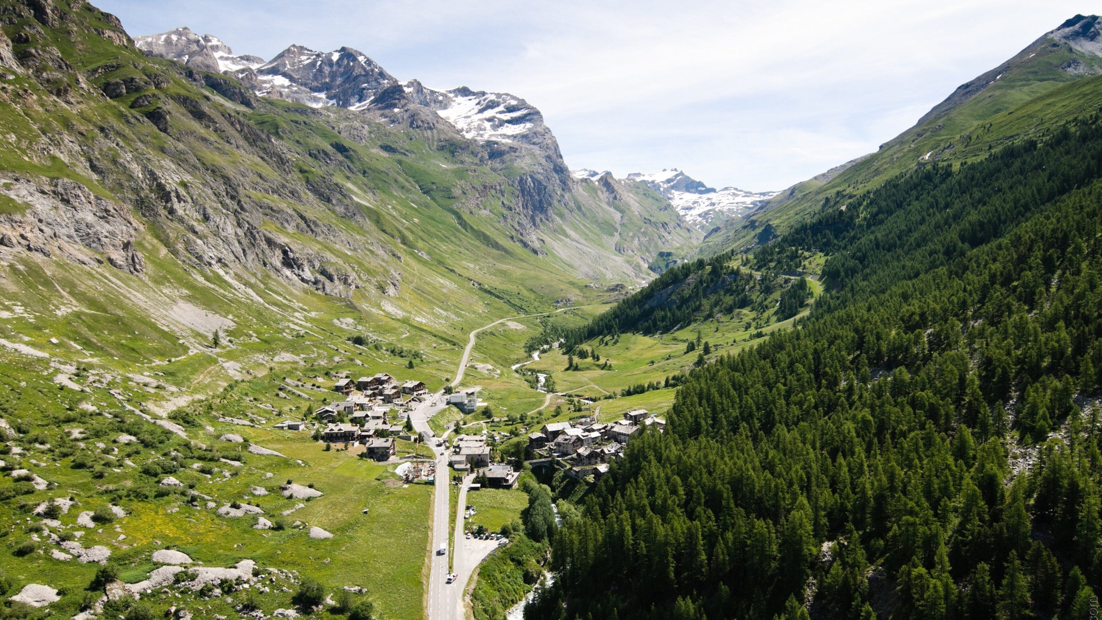 A mountain village nestled in a valley with a winding road, surrounded by lush greenery and rocky mountains.