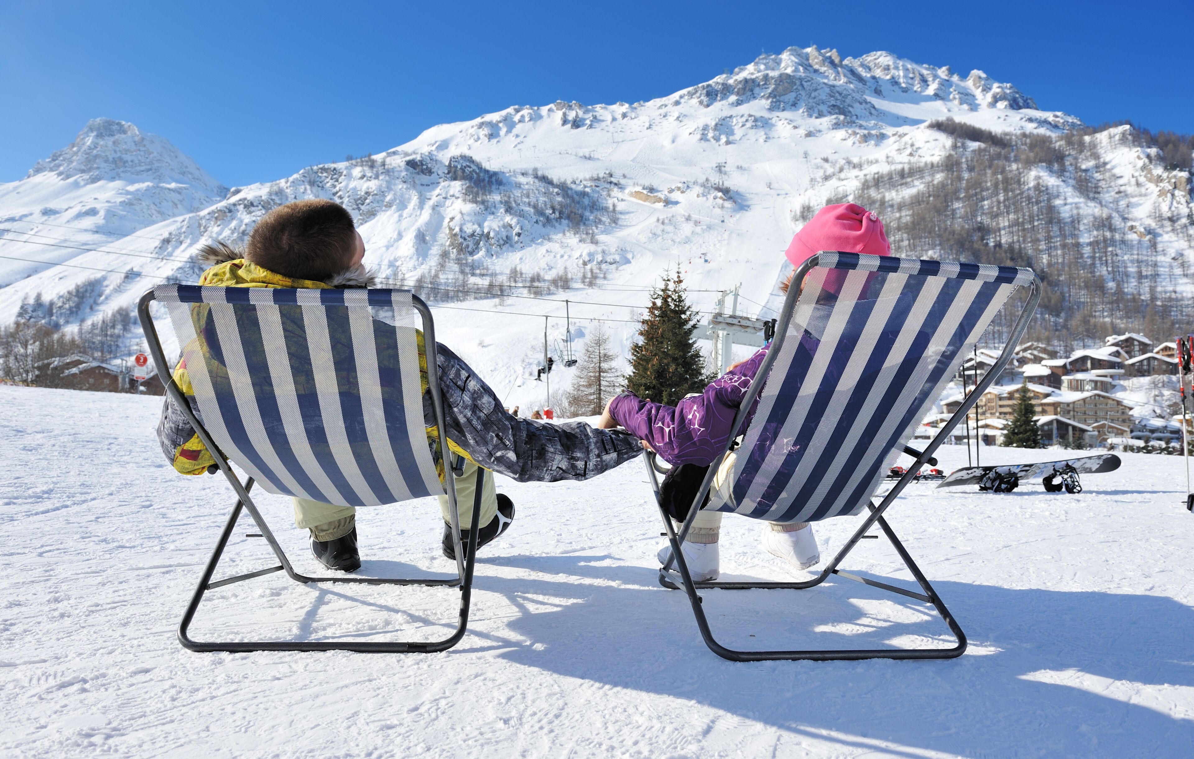 Dos personas sentadas en sillas de playa a rayas frente a un paisaje montañoso nevado con telesillas al fondo.