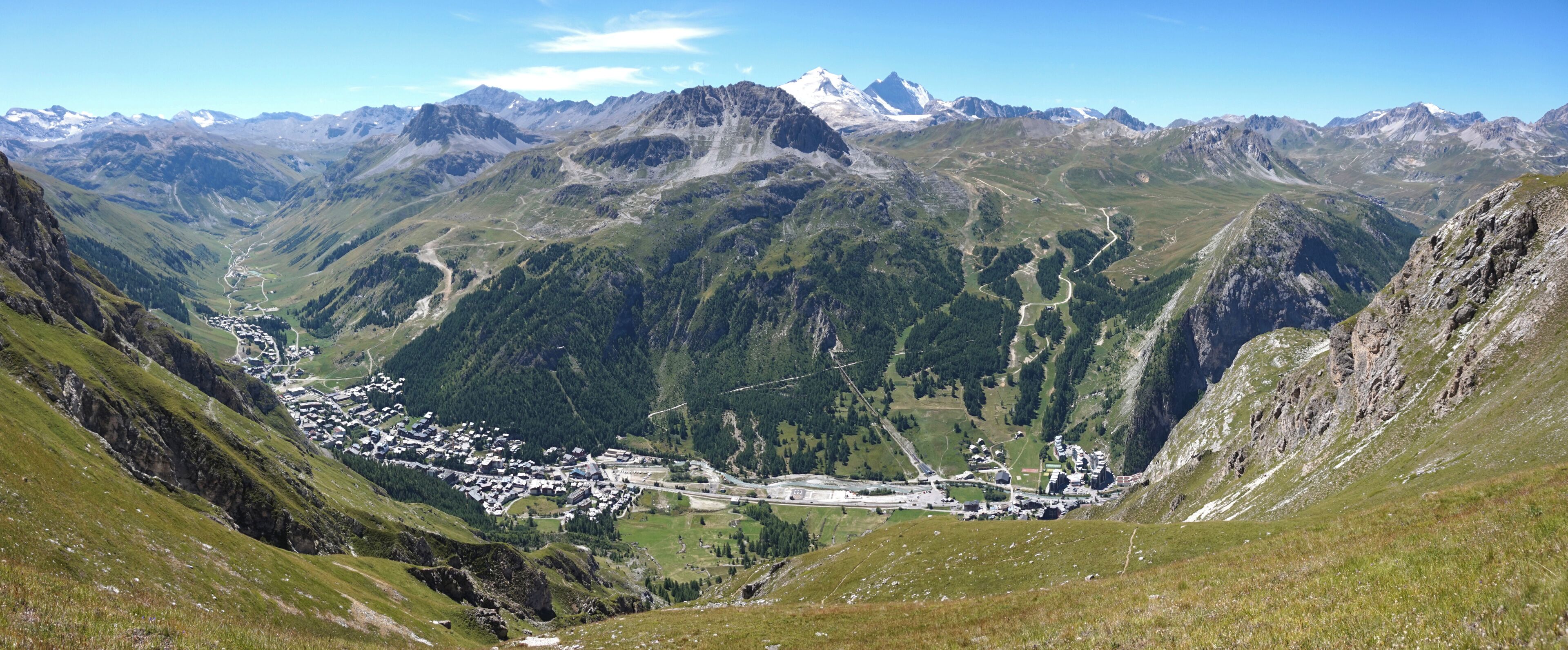 A valley with a town nestled between mountains, a winding road, and a snow-capped peak in the distance.