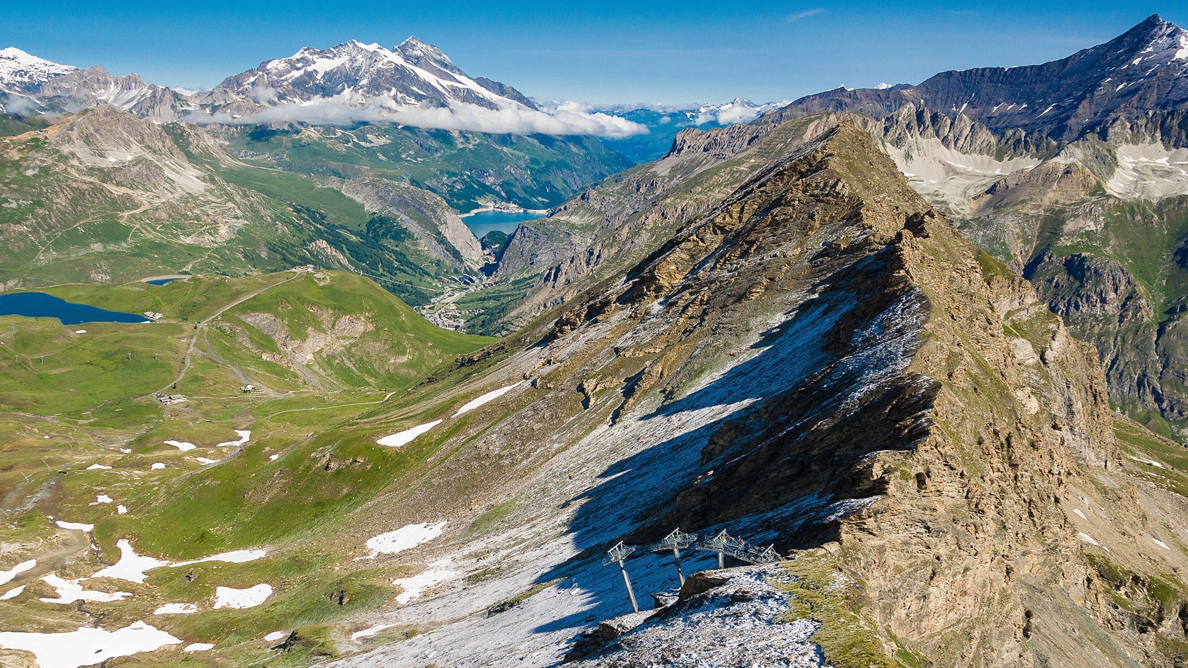 A mountainous landscape with snow-capped peaks, a valley with a lake, and a winding road.