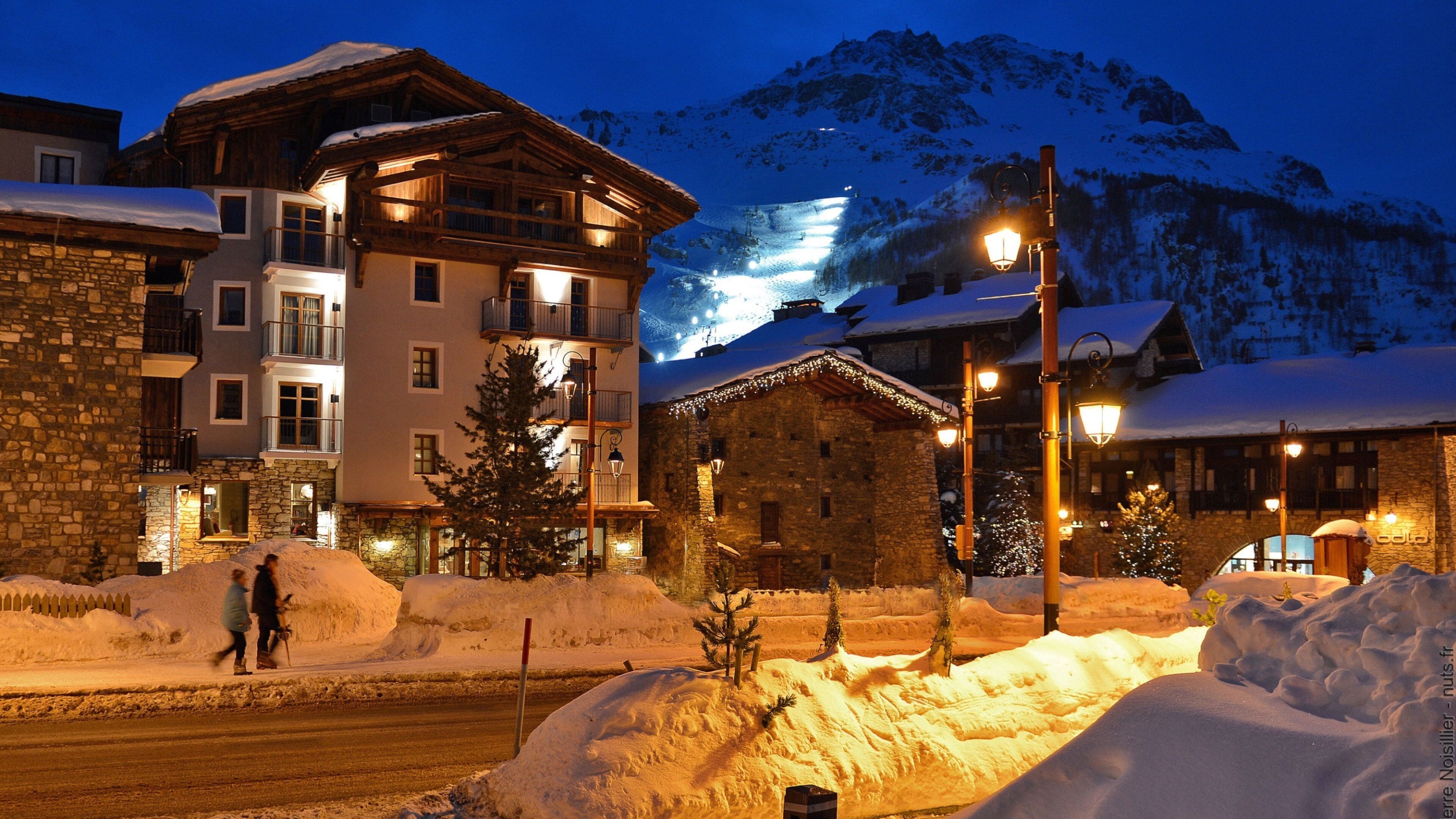 A snow-covered mountain at dusk with illuminated buildings and a wooden structure in the foreground.