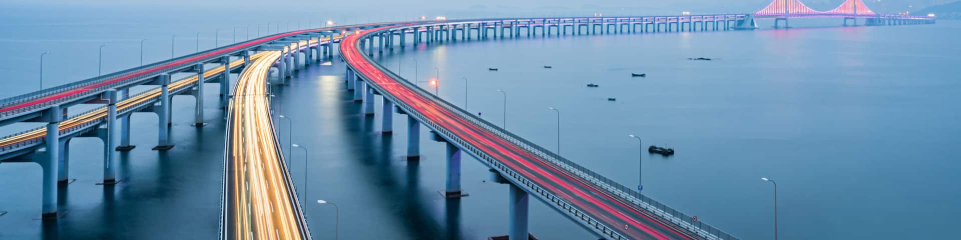 A long bridge with light trails, a calm sea, and distant red-lit structures.