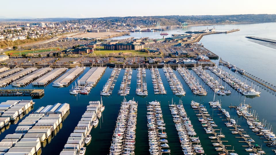 A marina with numerous boats docked, a large industrial area in the background, and a clear view of the waterfront.