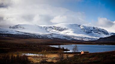 A snow-covered mountain range, a lake, and a small building in a valley.