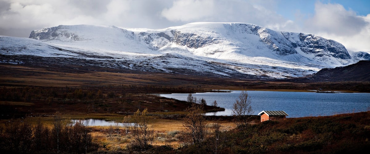 A snow-covered mountain range, a lake, and a small building in a valley.