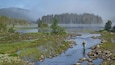A person fishing in a calm river surrounded by rocks and greenery.