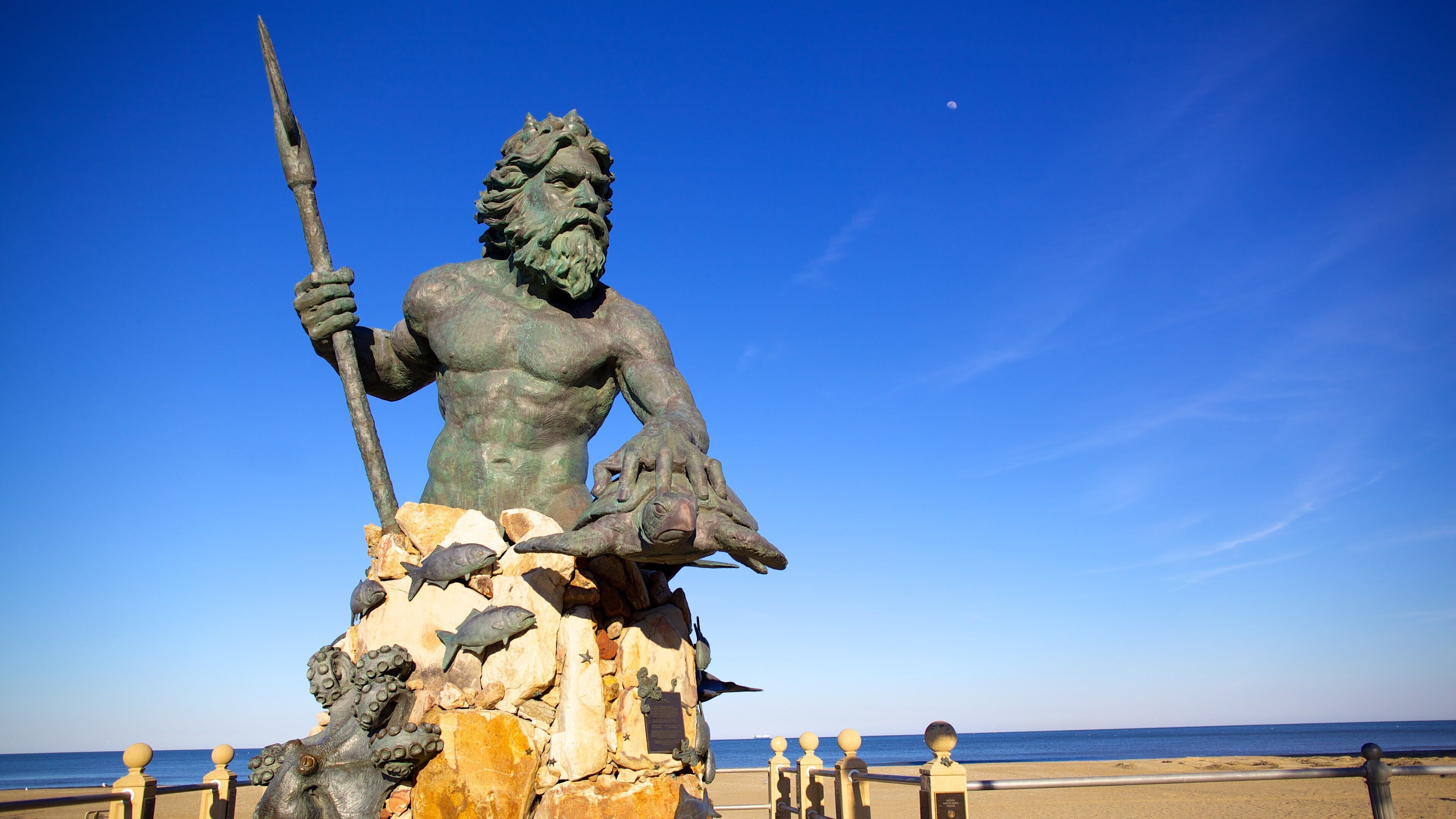 A bronze statue of a muscular figure holding a spear, seated on a rock with fish and sea creatures around it, overlooking a beach and the sea.