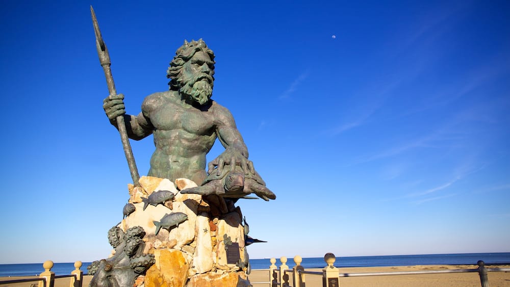 Une statue de bronze représentant une figure musclée tenant une lance, assise sur un rocher entouré de poissons et d’autres créatures marines, et dominant une plage et la mer.