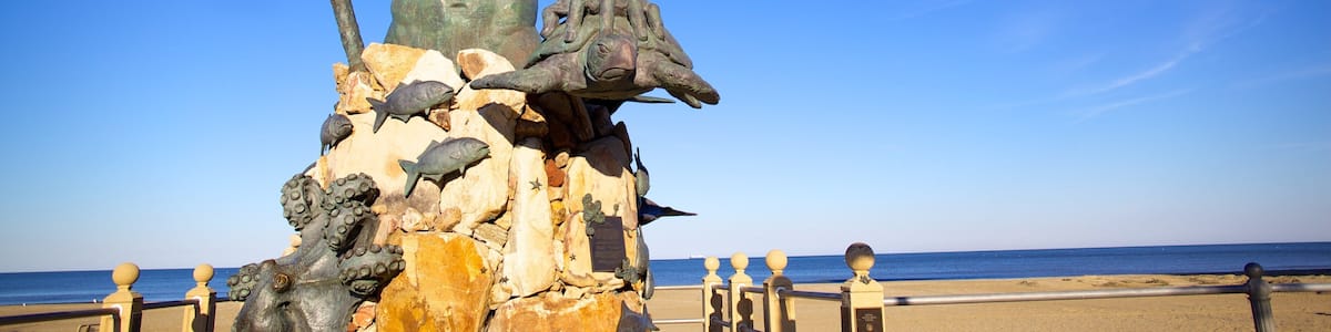 Une statue de bronze représentant une figure musclée tenant une lance, assise sur un rocher entouré de poissons et d’autres créatures marines, et dominant une plage et la mer.