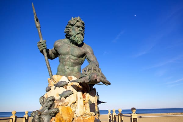 A bronze statue of a muscular figure holding a spear, seated on a rock with fish and sea creatures around it, overlooking a beach and the sea.
