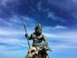 Una estatua de una figura musculosa sosteniendo una lanza, rodeada de rocas y peces, contra un cielo azul con nubes.
